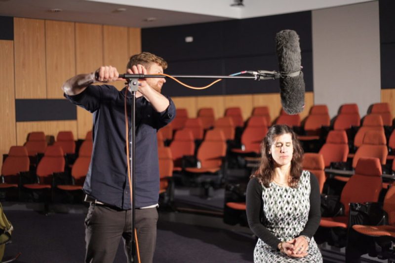 Zel Iscel is seated with sound man adjusting a boom microphone above her head. 