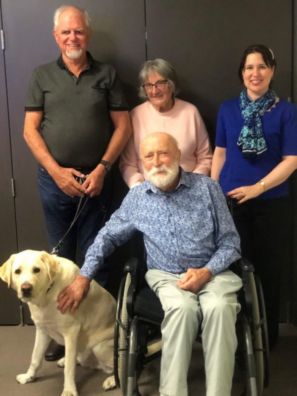 Four people in a group. One person is in a wheel chair. A man standing up has a Guide Dog.