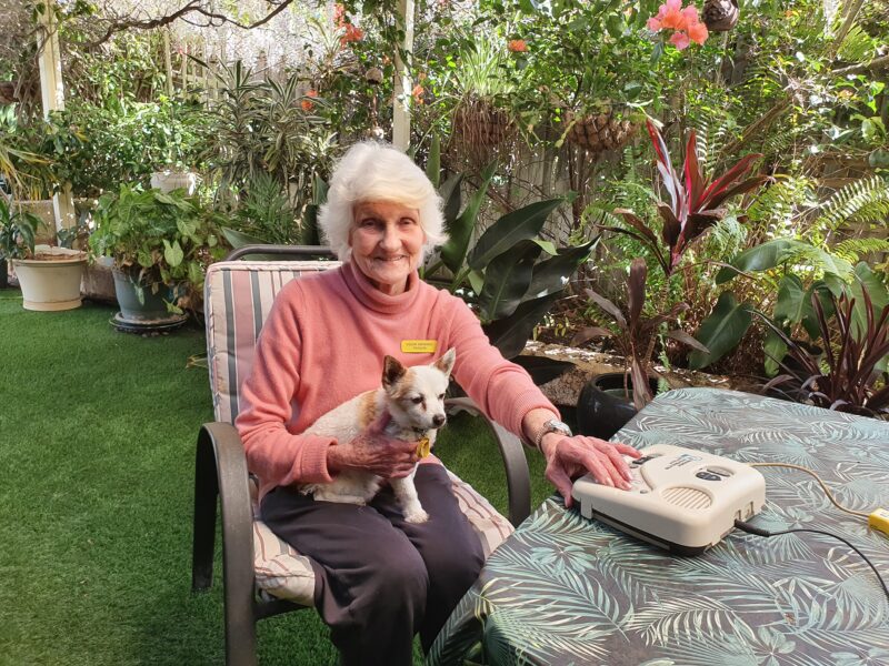 Hilary sits in a garden around a table with dog on her lap and listening to audio books 