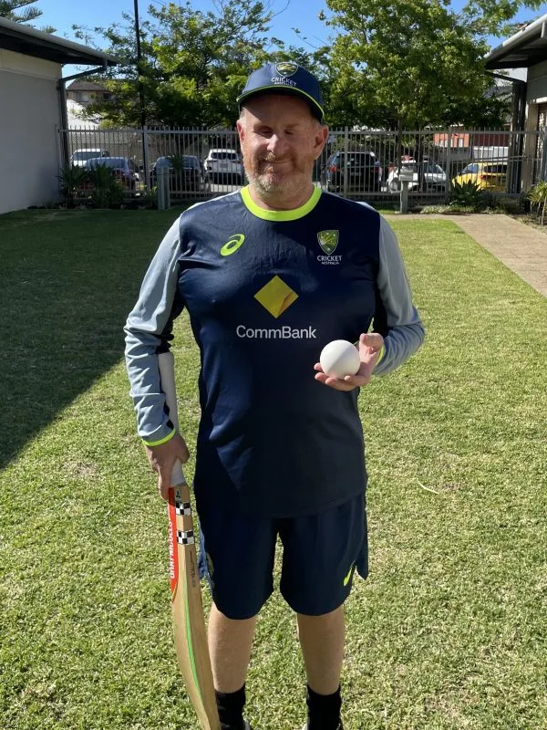 Ryan stands outside wearing his Australia uniform and holding a cricket bat and ball.